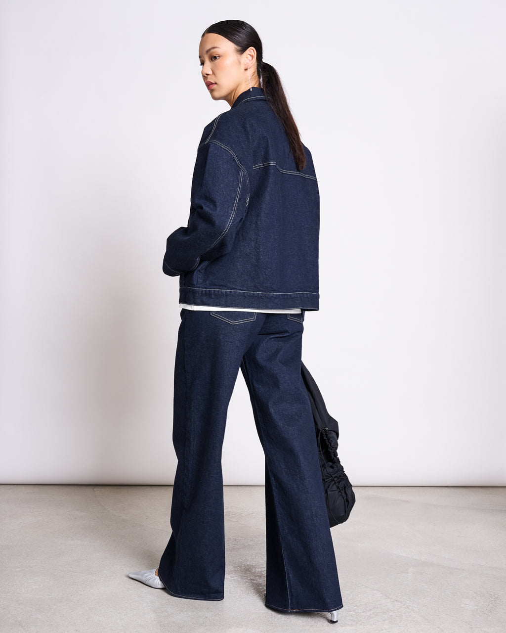 A woman with long dark hair in a low ponytail wears the jan-n-june RELAXED DENIM JACKET VILME DARK BLUE DENIM GOTS and wide-leg jeans, holding a black bag in a minimalist indoor space with a light background.