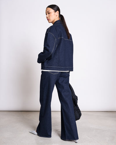 A woman with long dark hair in a low ponytail wears the jan-n-june RELAXED DENIM JACKET VILME DARK BLUE DENIM GOTS and wide-leg jeans, holding a black bag in a minimalist indoor space with a light background.