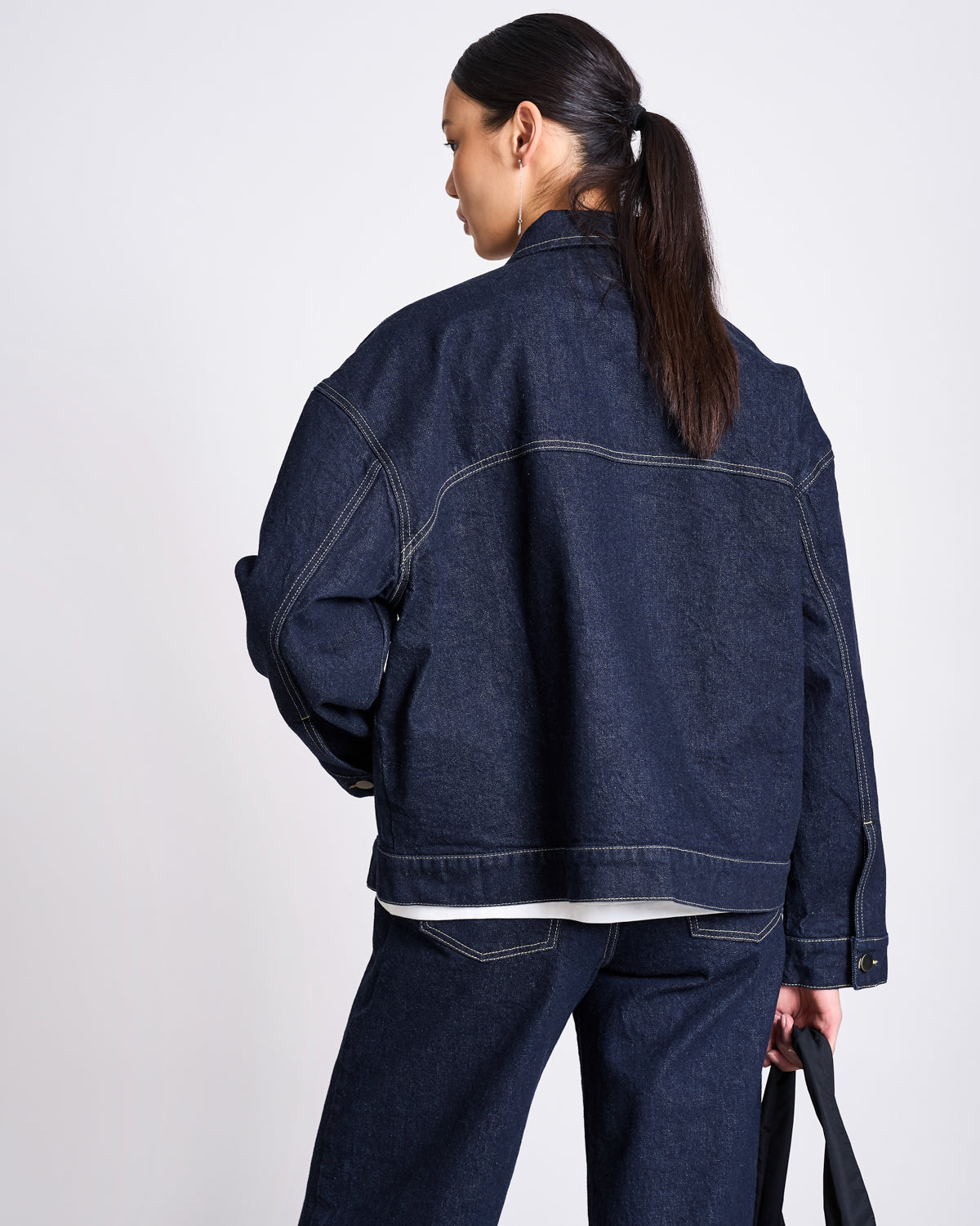 A woman with long dark hair in a low ponytail wears the jan-n-june RELAXED DENIM JACKET VILME DARK BLUE DENIM GOTS and matching jeans. With her back to the camera, she holds a black bag in her left hand.