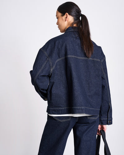 A woman with long dark hair in a low ponytail wears the jan-n-june RELAXED DENIM JACKET VILME DARK BLUE DENIM GOTS and matching jeans. With her back to the camera, she holds a black bag in her left hand.