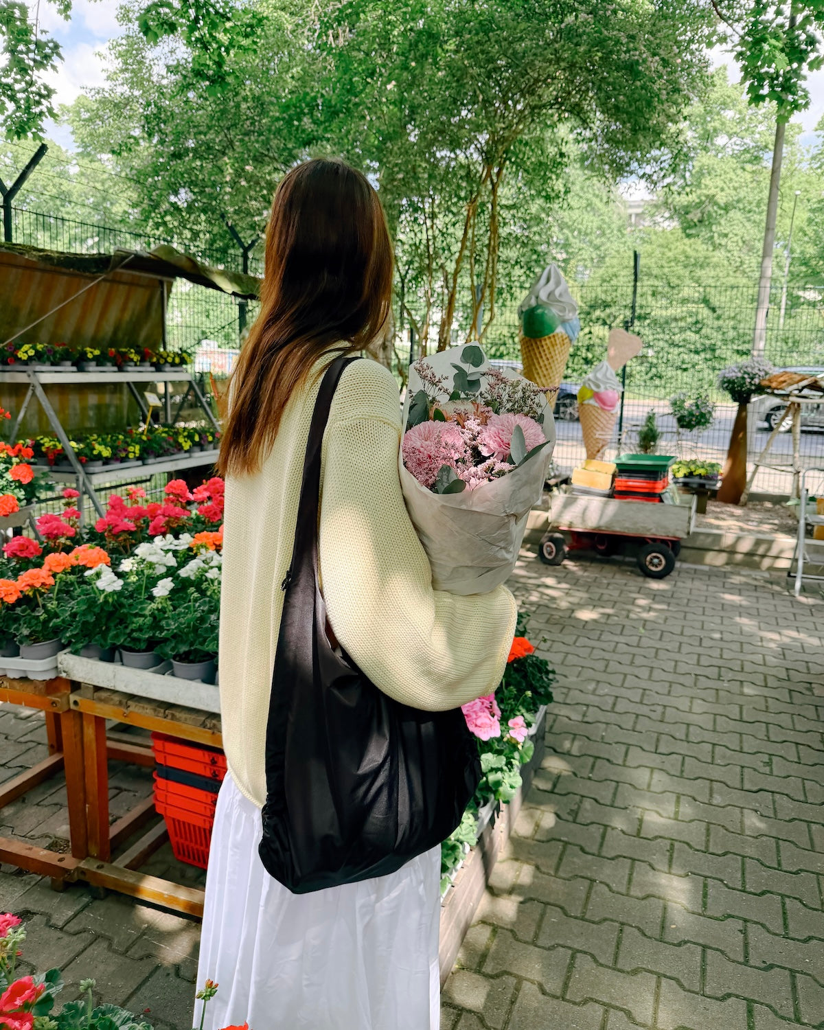 A woman with long brown hair wears the jan-n-june JUMPER SONA BUTTERMILK GOTS sweater and a white skirt, standing outside at a market with colorful potted flowers, holding pink flowers and a black bag.