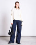 A woman stands against a plain background wearing jan-n-june’s LIGHT KNIT JUMPER WILLIAMSBURG OFFWHITE GOTS, wide-leg dark blue jeans, and black shoes. She holds a black shoulder bag and wears a chunky silver necklace.