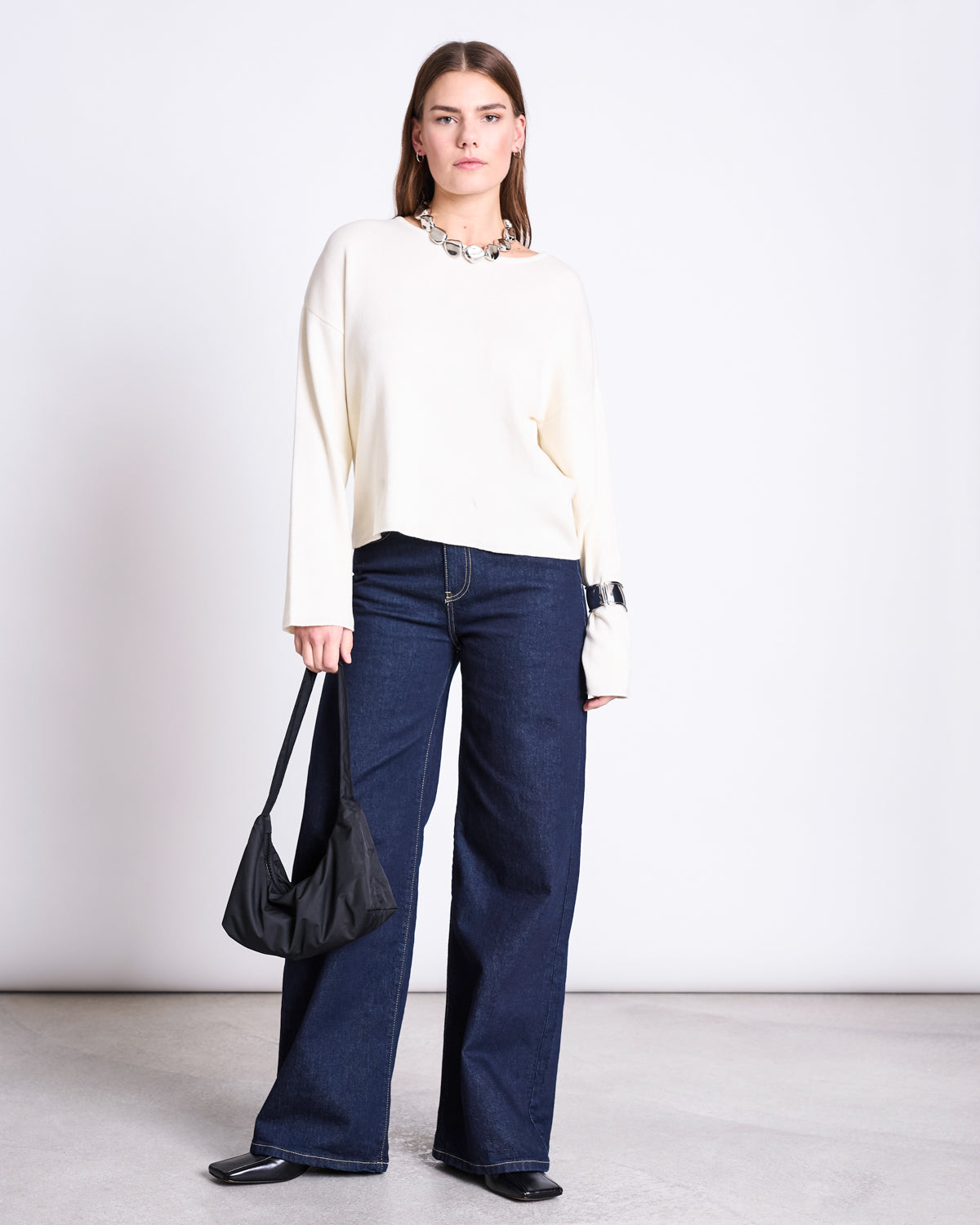 A woman stands against a plain background wearing jan-n-june’s LIGHT KNIT JUMPER WILLIAMSBURG OFFWHITE GOTS, wide-leg dark blue jeans, and black shoes. She holds a black shoulder bag and wears a chunky silver necklace.