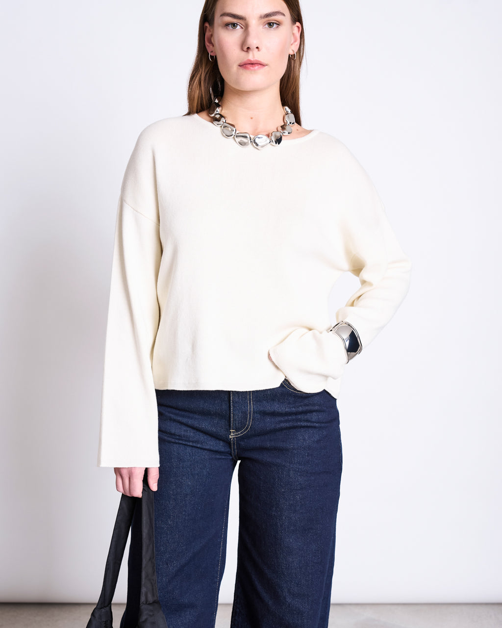 A woman poses against a plain background in the jan-n-june LIGHT KNIT JUMPER WILLIAMSBURG OFFWHITE GOTS, paired with dark blue jeans and a chunky silver necklace. One hand rests on her hip while a black strap drapes over her other arm.