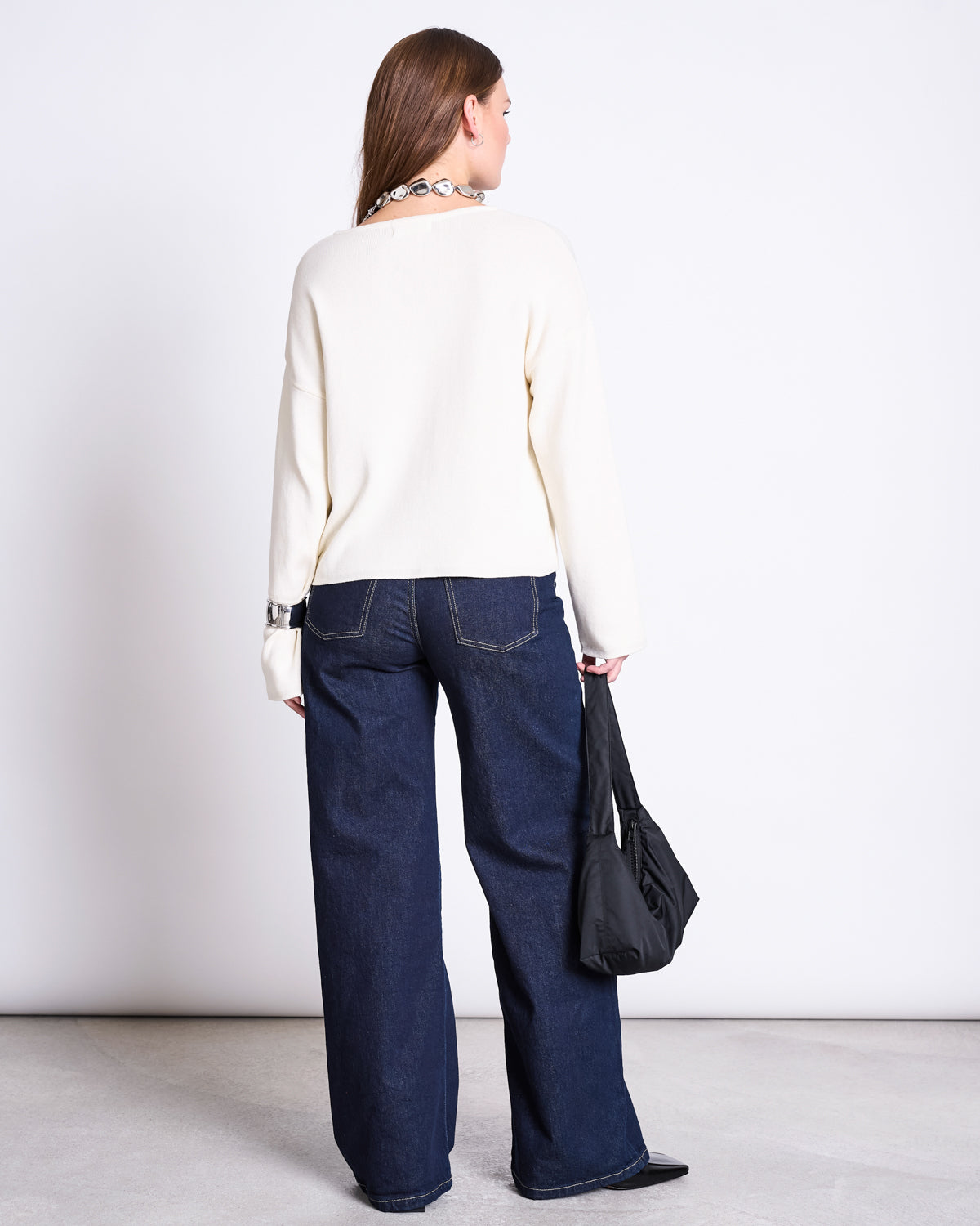 A woman faces away, wearing the jan-n-june LIGHT KNIT JUMPER WILLIAMSBURG OFFWHITE GOTS and dark wide-leg jeans with visible stitching. She holds a black handbag; her straight brown hair stands out against a plain white background.