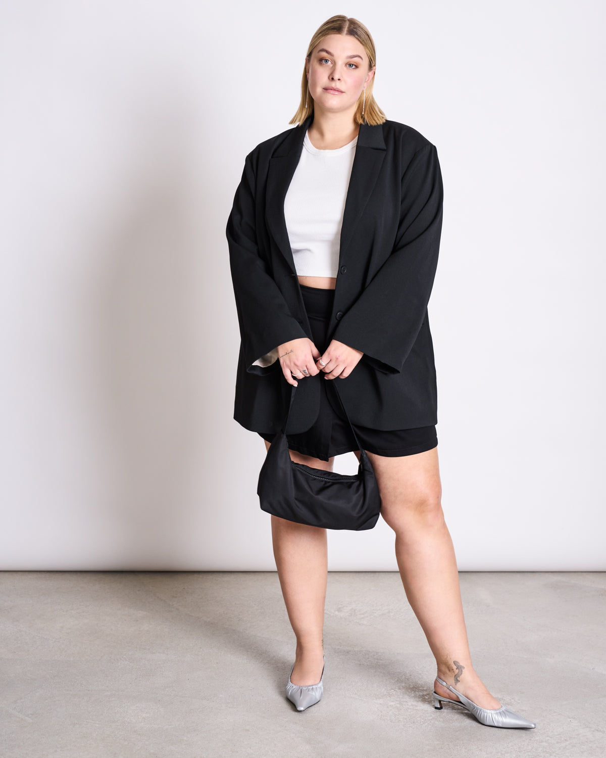 A woman with blond hair wears a white crop top, black shorts, silver heels, and the jan-n-june BLAZER DURANCE BLACK made from recycled polyester. She holds a black handbag and stands against a plain white background.