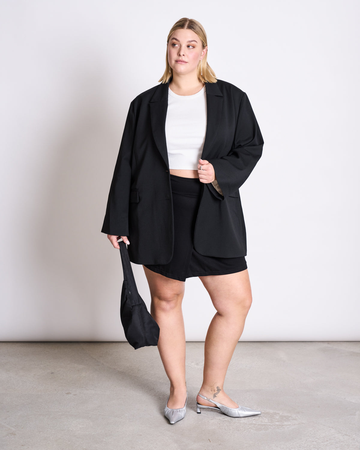 A woman stands on a concrete floor holding a black handbag, wearing jan-n-junes BLAZER DURANCE BLACK over a white crop top, black mini skirt, and silver low-heeled shoes, looking to the side against a plain white background.