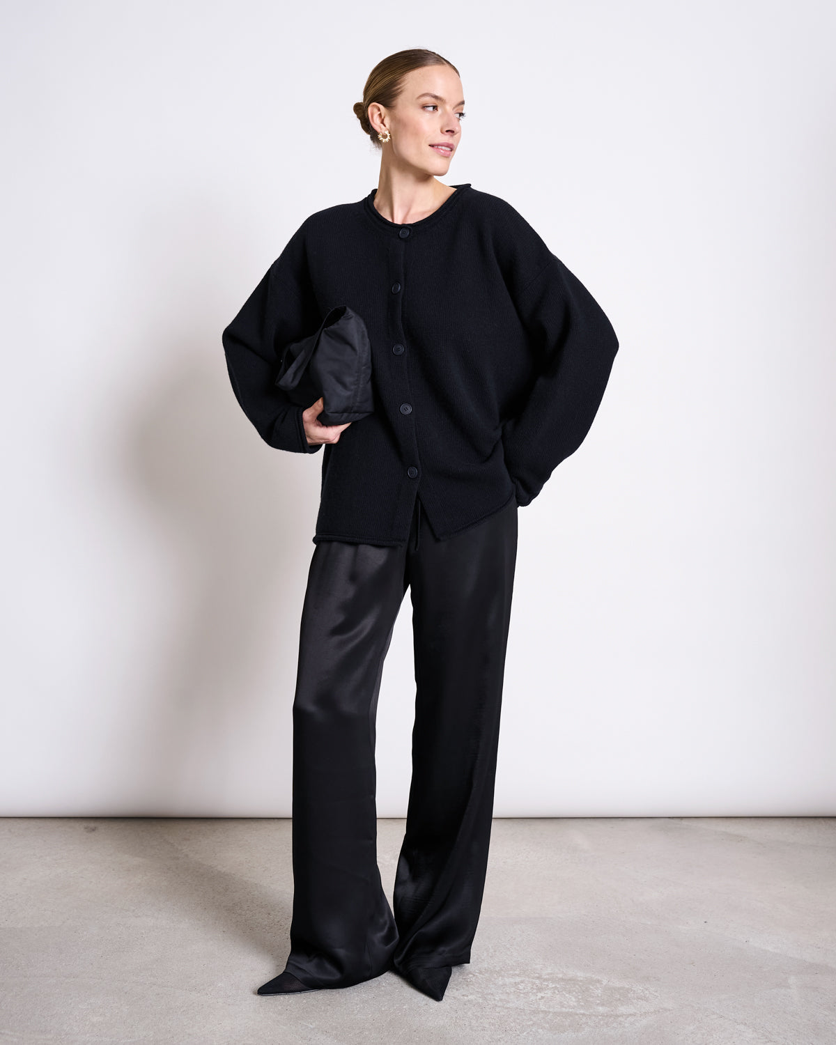 A woman stands against a white wall, wearing the jan-n-june OVERSIZED MERINO WOOL CARDIGAN TINCO BLACK GOTS and wide-leg black satin pants, holding a black clutch. Her hair is pulled back as she smiles slightly, looking to the side.