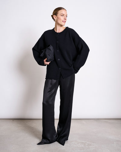 A woman stands against a white wall, wearing jan-n-june’s LOOSE SILKY PANTS FRIS BLACK with a flexible waistband, paired with a loose black cardigan and matching clutch. Her hair is pulled back as she looks to the side neutrally.