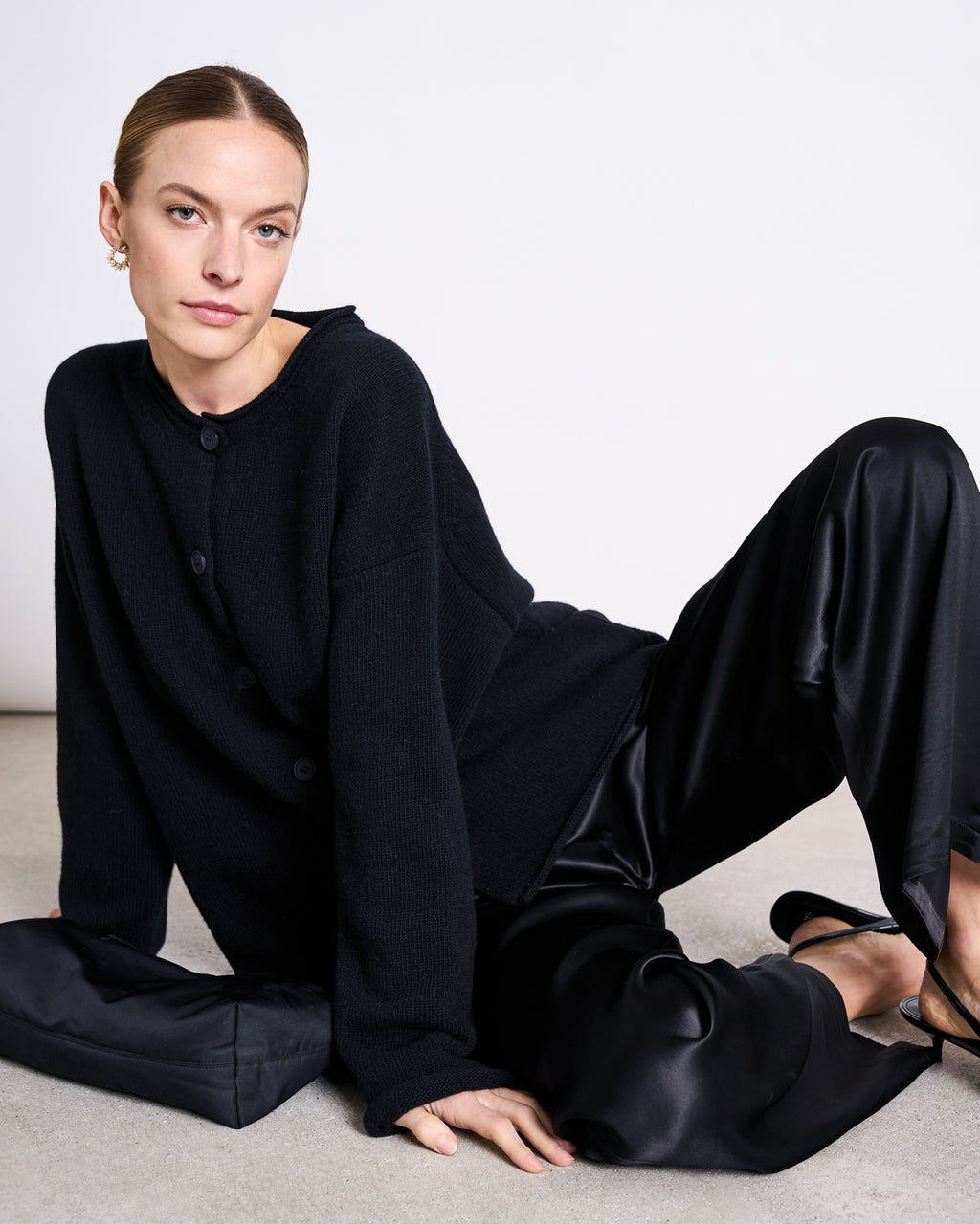 A woman with slicked-back hair sits on the floor against a white background, wearing the jan-n-june OVERSIZED MERINO WOOL CARDIGAN TINCO BLACK GOTS, black satin pants, and black heels, looking directly at the camera with a neutral expression.
