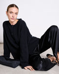 A woman with slicked-back hair sits on the floor against a white background, wearing the jan-n-june OVERSIZED MERINO WOOL CARDIGAN TINCO BLACK GOTS, black satin pants, and black heels, looking directly at the camera with a neutral expression.