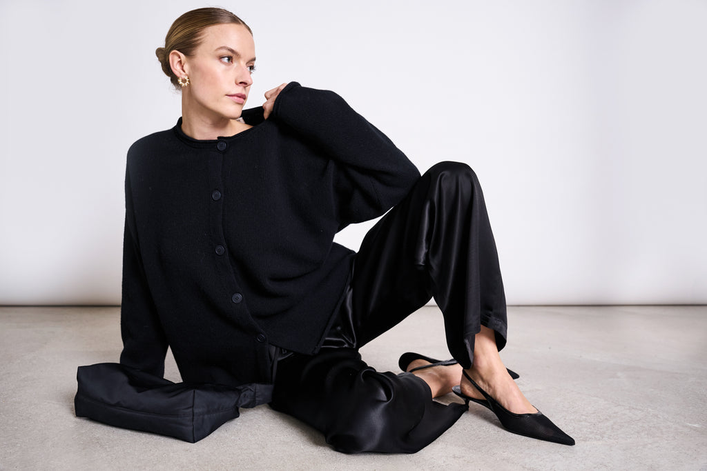 A woman wears the jan-n-june OVERSIZED MERINO WOOL CARDIGAN TINCO BLACK GOTS with black satin pants, black heels, gold earrings, and a black bag. She sits on the floor in a plain white setting, looking to the side with one leg bent.