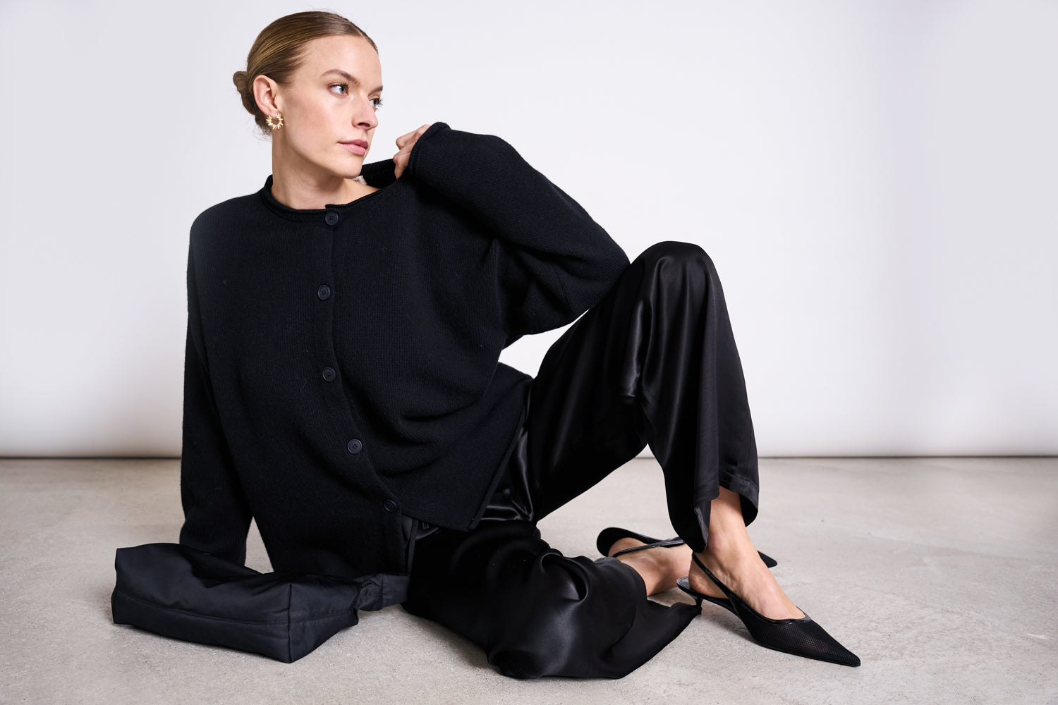 A woman with blonde hair in a bun wears the jan-n-june LOOSE SILKY PANTS FRIS BLACK with a black sweater and heels, sitting on a light floor next to a black clutch, looking left against a plain white background.