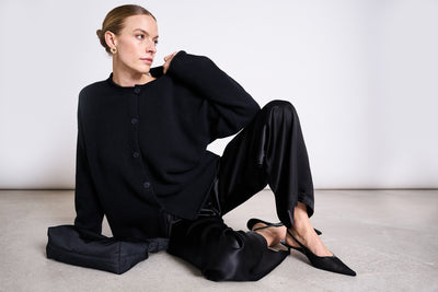 A woman with blonde hair in a bun wears the jan-n-june LOOSE SILKY PANTS FRIS BLACK with a black sweater and heels, sitting on a light floor next to a black clutch, looking left against a plain white background.