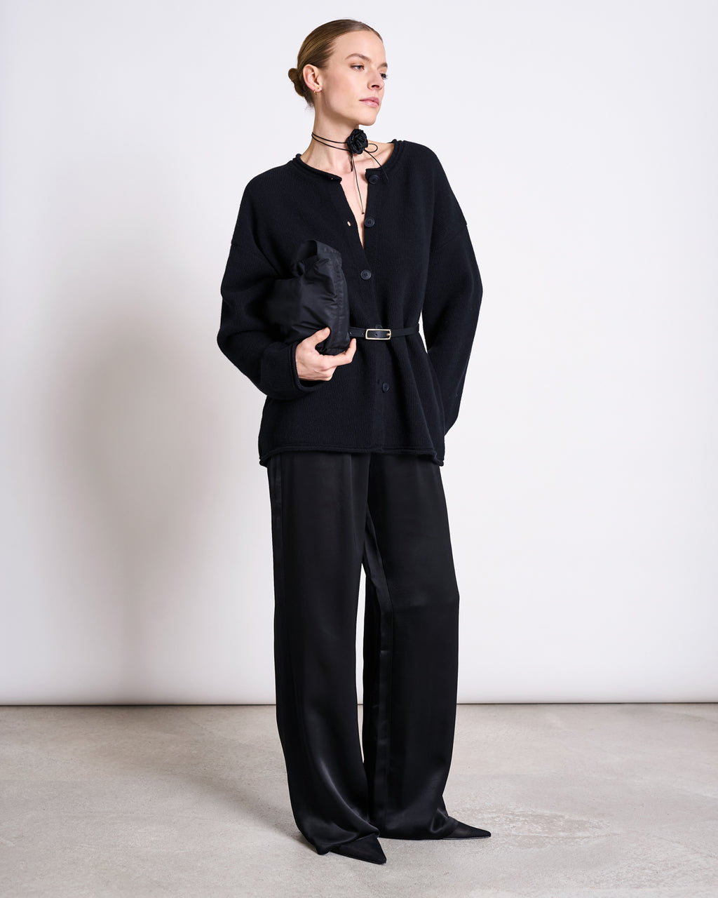 A person stands against a plain white wall, wearing the jan-n-june OVERSIZED MERINO WOOL CARDIGAN TINCO BLACK GOTS, wide-leg black pants, pointed black shoes, hair pulled back, and holding a folded black garment under one arm.