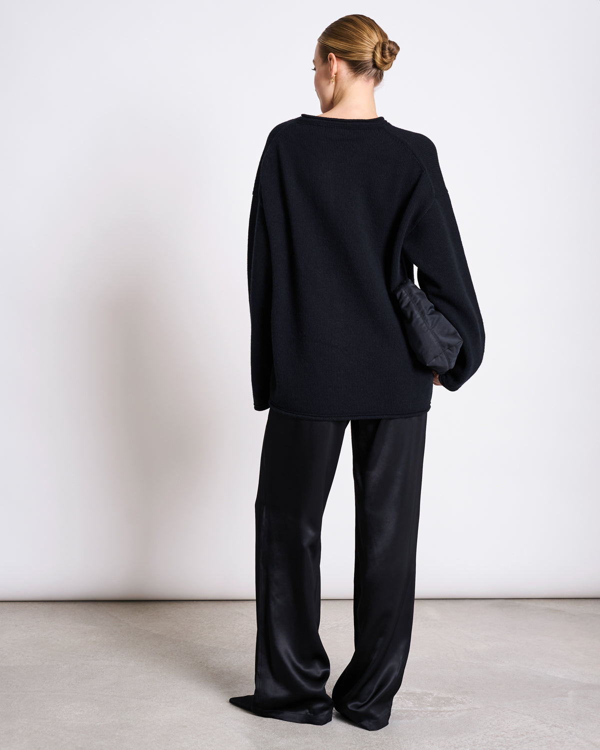 A woman with a neat bun stands with her back to the camera, wearing the jan-n-june OVERSIZED WOOL CARDIGAN TINCO BLACK GOTS, loose black satin pants, and holding a small black clutch against a minimalistic background.