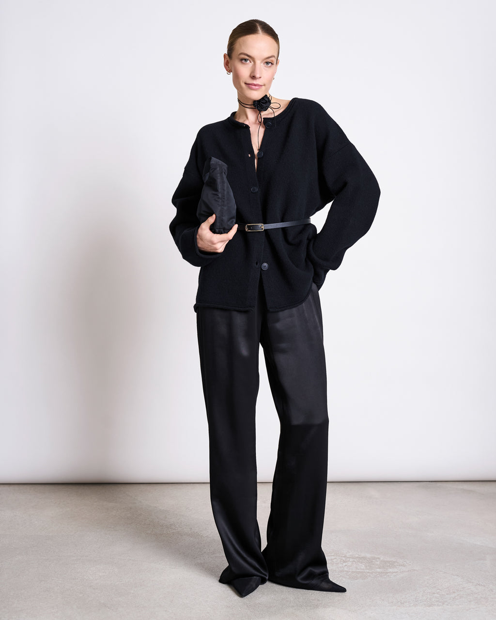A woman stands against a plain background wearing the jan-n-june OVERSIZED WOOL CARDIGAN TINCO BLACK GOTS, styled with black wide-leg satin pants, a slim black belt, black sandals, and a matching clutch, looking confidently at the camera.
