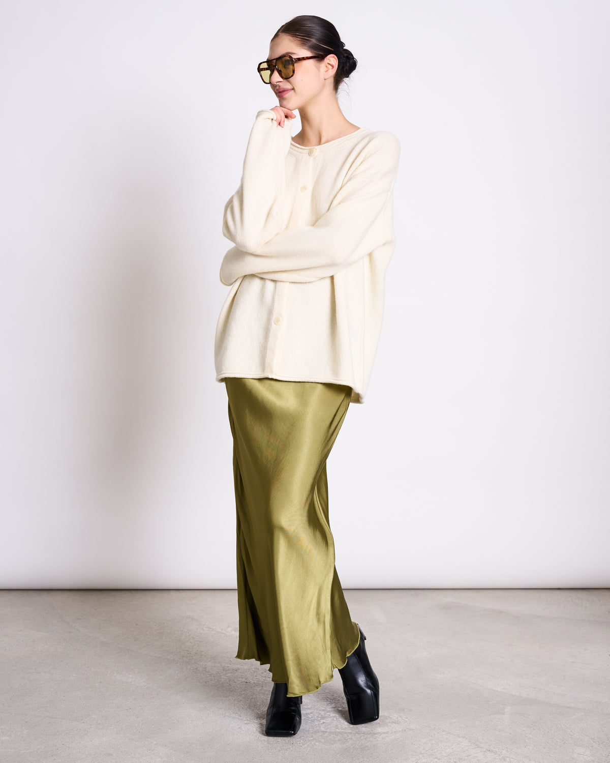 A woman in sunglasses and a bun wears the jan-n-june OVERSIZED MERINO WOOL CARDIGAN TINCO OFFWHITE GOTS over an olive green satin skirt, black platform shoes, standing against a plain backdrop with one arm folded, looking sideways.