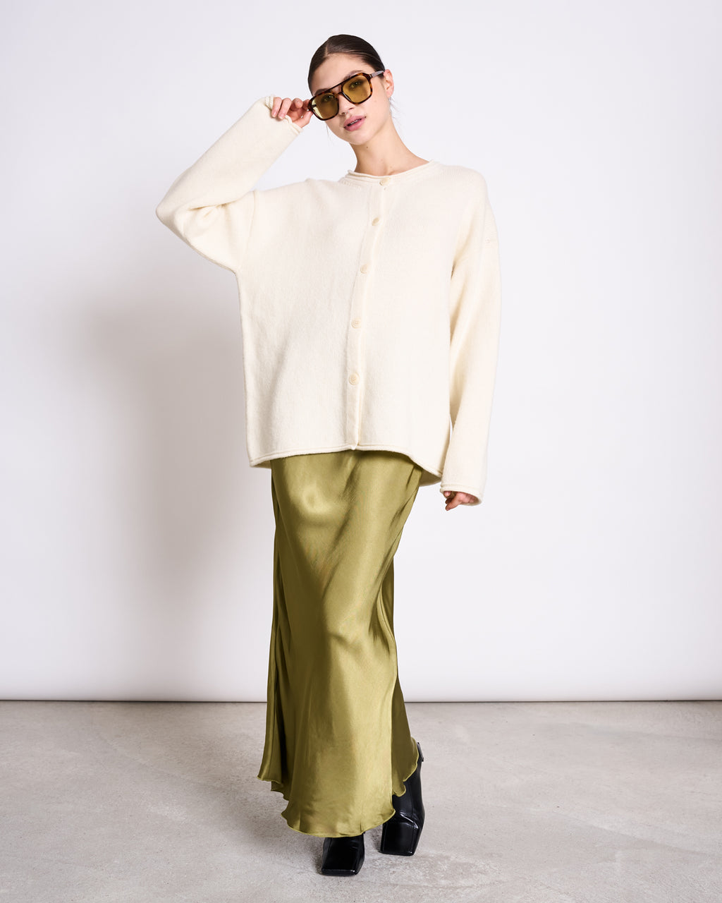 A woman in oversized sunglasses and the jan-n-june OVERSIZED MERINO WOOL CARDIGAN TINCO OFFWHITE GOTS stands on a light-gray floor by a white wall, wearing a long olive satin skirt and posing with one hand on her sunglasses.