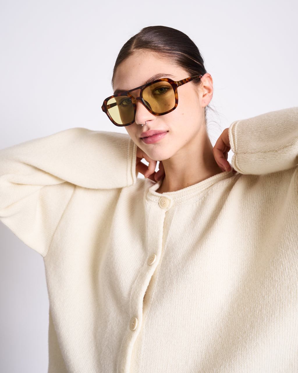 A woman poses confidently with her hands behind her neck, wearing the jan-n-june OVERSIZED MERINO WOOL CARDIGAN TINCO OFFWHITE GOTS and yellow-tinted sunglasses against a plain background.