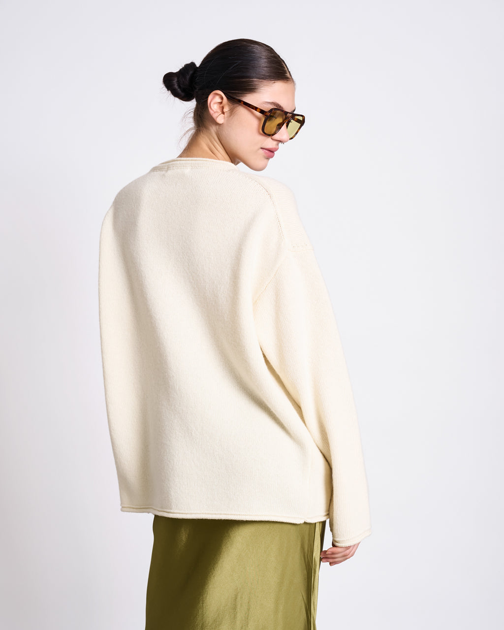 A person with dark hair in a bun wears yellow-tinted sunglasses and the jan-n-june OVERSIZED WOOL CARDIGAN TINCO OFFWHITE GOTS with an olive green skirt, standing against a plain white background, looking over their shoulder.