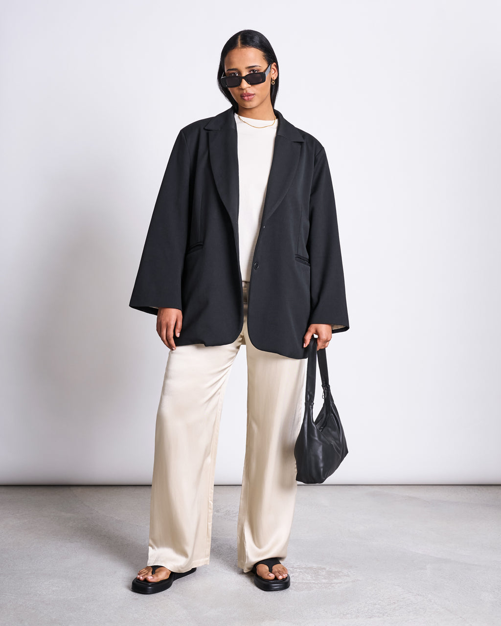 A person stands against a plain background wearing sunglasses, a black oversized blazer, black sandals, and holding a black handbag. They are styled in jan-n-june LOOSE SILKY PANTS FRIS IVORY with a low-waist fit.