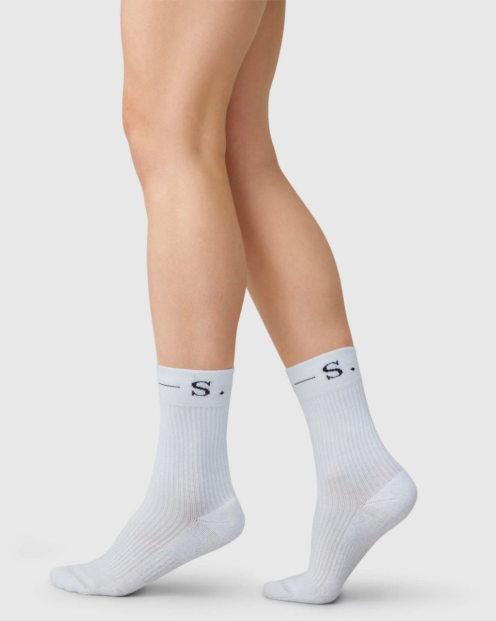 A person stands on tiptoes against a light background, wearing jan-n-june SWEDISH STOCKINGS SOCKS BELLA WHITE—classic sport socks made from durable organic cotton, featuring ribbed texture and black “S.” lettering near the top.