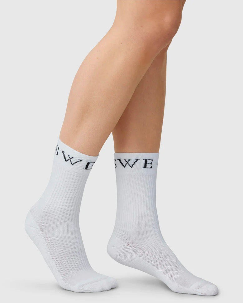 A person wearing jan-n-june SWEDISH STOCKINGS SOCKS BELLA WHITE, classic organic cotton crew socks with white ribbed design and black lettering near the top, stands on a plain light background. Only their lower legs and feet are visible.