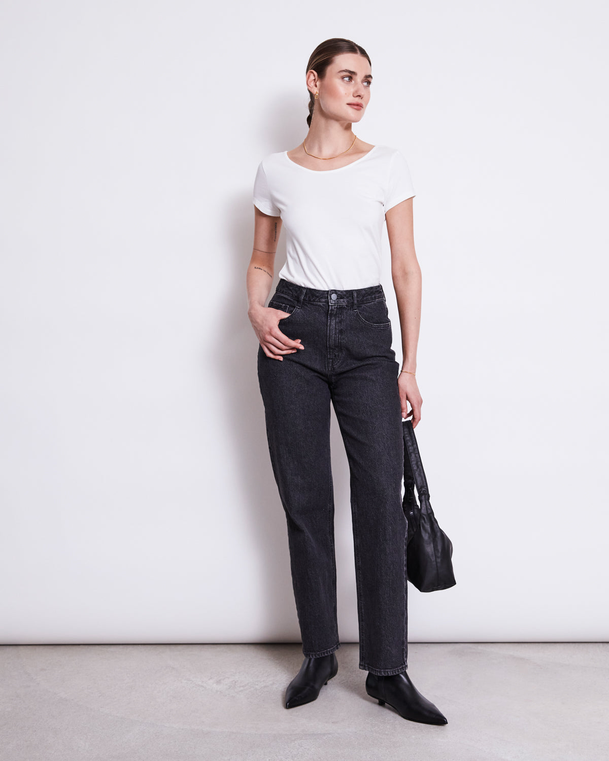 A woman stands against a white wall wearing the jan-n-june BALLERINA T-SHIRT ALOE WHITE GOTS, paired with high-waisted black jeans and black shoes. She holds a black handbag and looks to the side, her hair tied back.