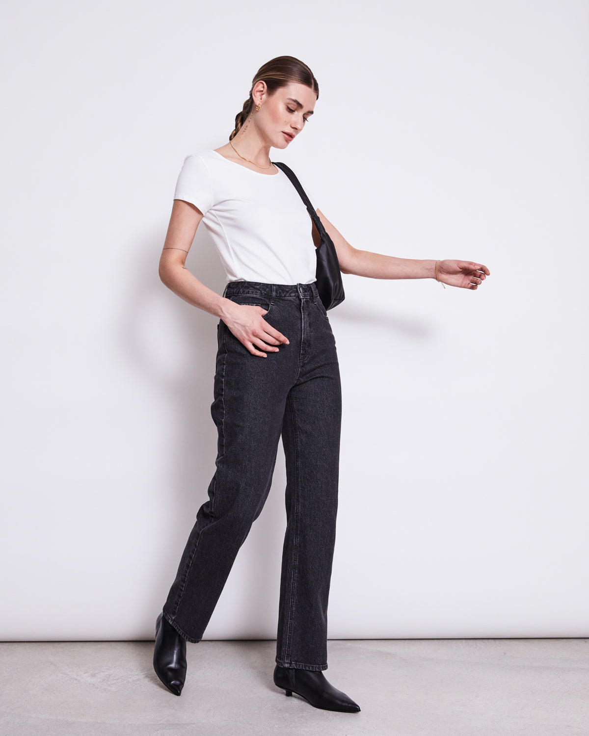 A woman stands against a white wall, wearing the jan-n-june BALLERINA T-SHIRT ALOE WHITE GOTS, high-waisted black jeans, black ankle boots, and a black shoulder bag, facing sideways with one arm extended.