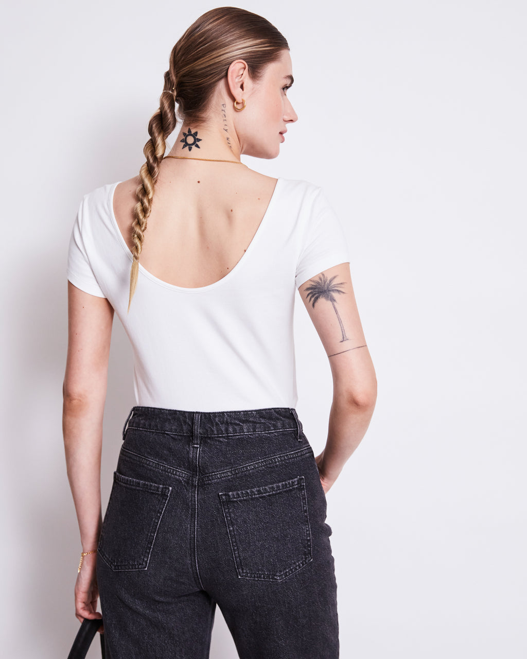 A woman with a long braid wears the jan-n-june BALLERINA T-SHIRT ALOE WHITE GOTS and black high-waisted jeans, revealing a sun neck tattoo and palm tree arm tattoo as she faces away against a plain white background.