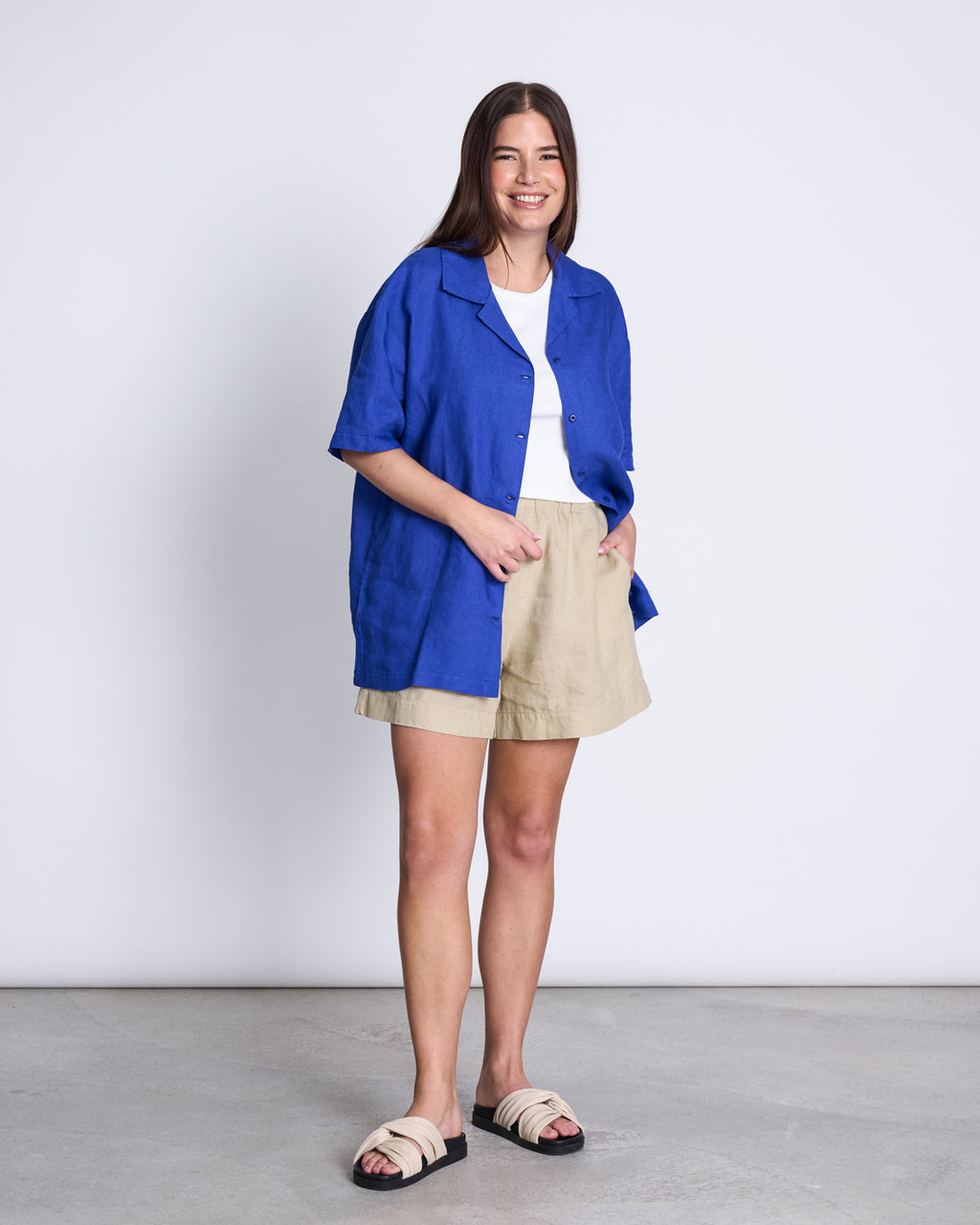 A woman smiles against a white background, wearing the jan-n-june LINEN SHIRT GENT BLUE over a white t-shirt, paired with beige shorts and beige slide sandals.