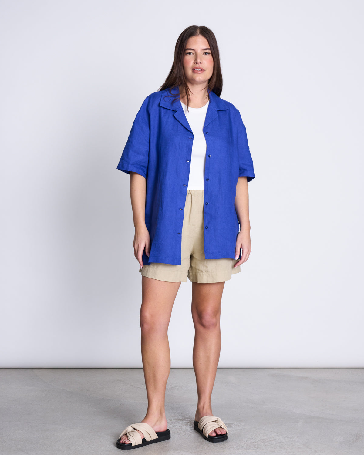A woman stands indoors against a white wall, wearing the jan-n-june LINEN SHIRT GENT BLUE over a white top with beige shorts and slide sandals. Her long brown hair and relaxed style complete this easy summer look.