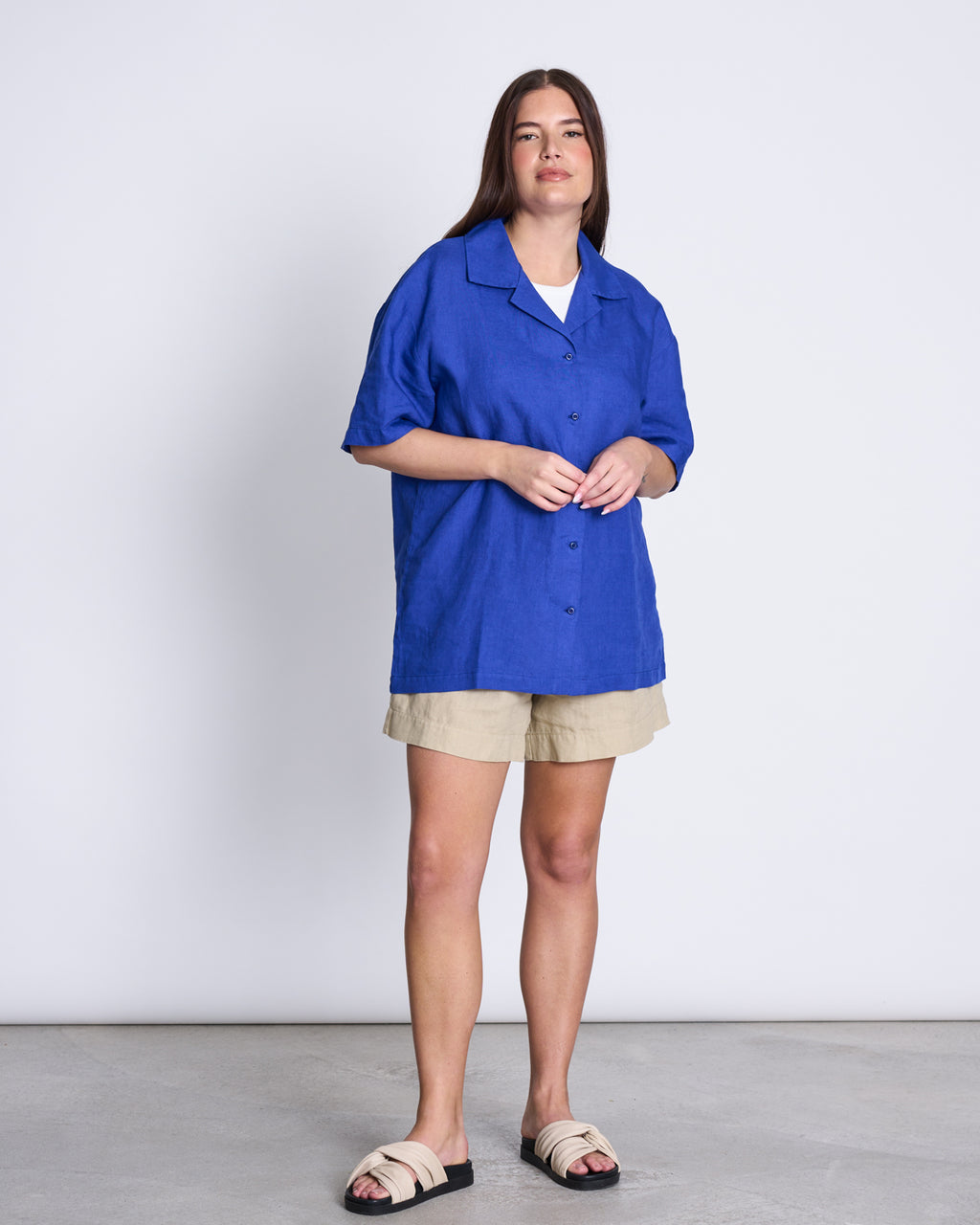 A woman with long brown hair stands against a white wall wearing the loose-fit jan-n-june LINEN SHIRT GENT BLUE, beige shorts, and beige slides for a relaxed, effortlessly unisex look.