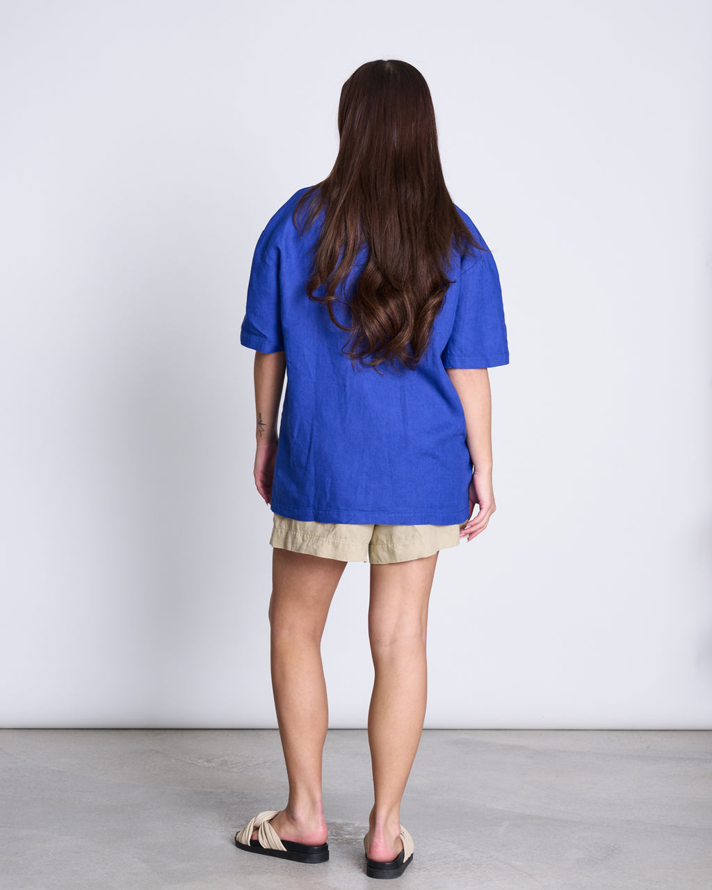 A woman with long brown hair, seen from behind, wears the jan-n-june LINEN SHIRT GENT BLUE paired with beige shorts and light sandals, standing against a plain white background.