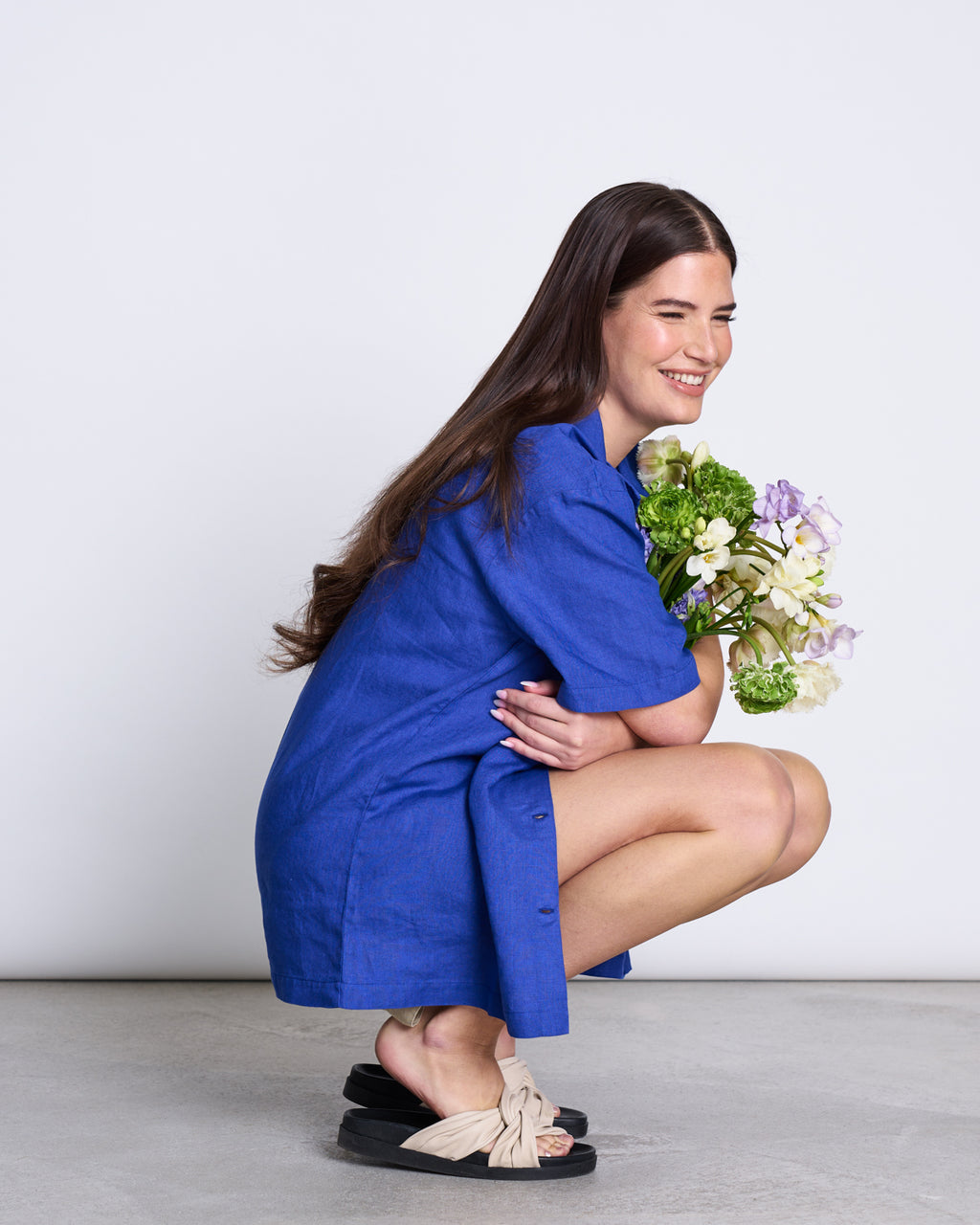 A woman with long brown hair smiles as she crouches, holding a small flower bouquet. Shes wearing jan-n-june’s LINEN SHIRT GENT BLUE as a dress with cream sandals, set against a plain white background.