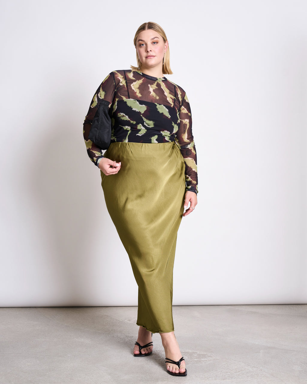 A woman poses against a plain background wearing the jan-n-june MESH LONGSLEEVE DORA MOSSFIRE, styled with an olive green satin skirt, black sandals, and a small black bag.