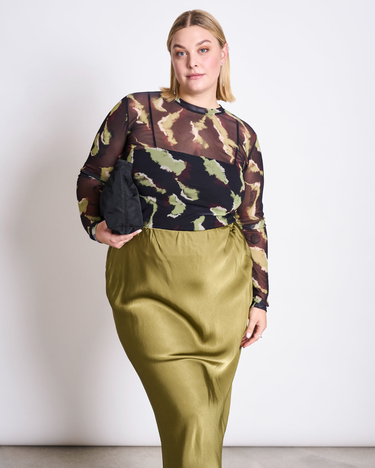 A blond-haired woman wears the jan-n-june MESH LONGSLEEVE DORA MOSSFIRE— a camouflage mesh top over black layers with a shiny olive-green skirt— and holds a black clutch against a white background.