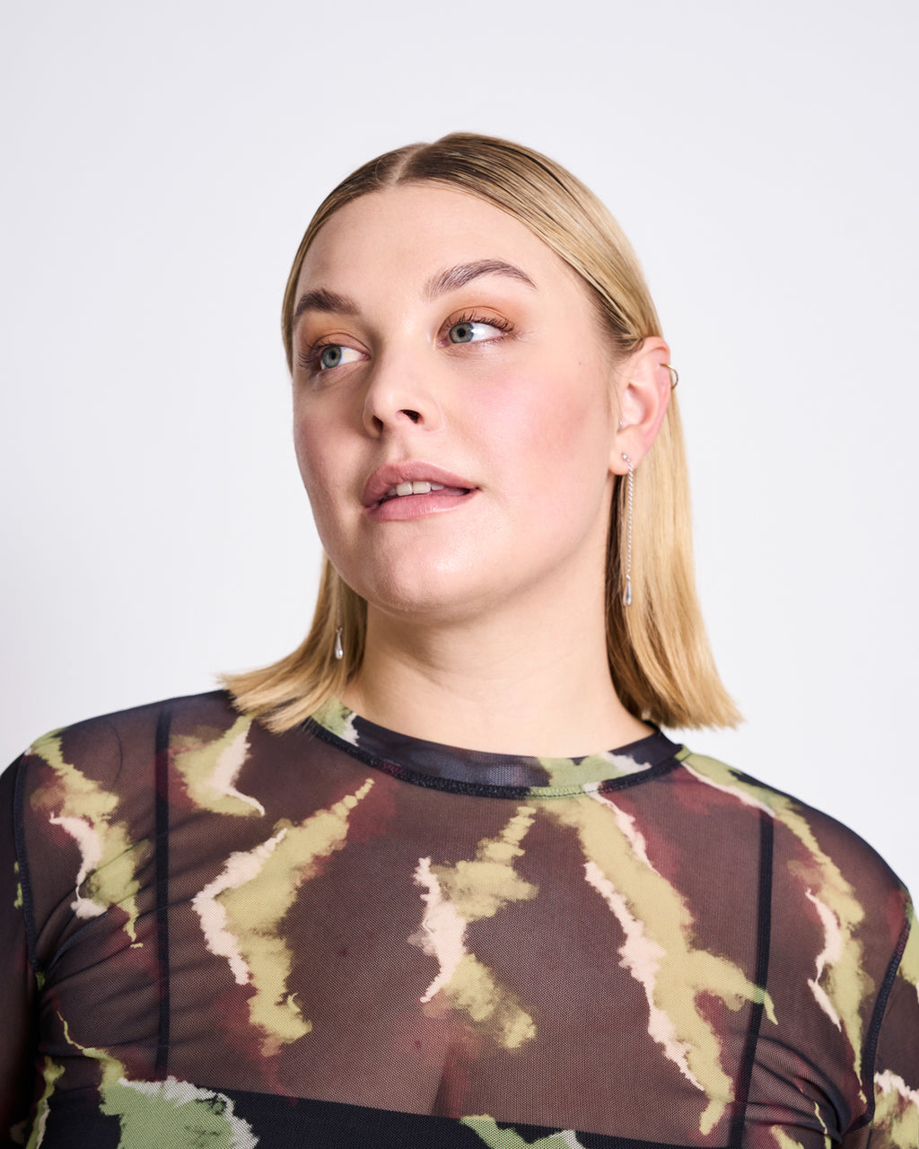 A woman with straight, shoulder-length blonde hair models the jan-n-june MESH LONGSLEEVE DORA MOSSFIRE in green, brown, and beige. She wears light makeup and long earrings, gazing slightly left against a plain light background.