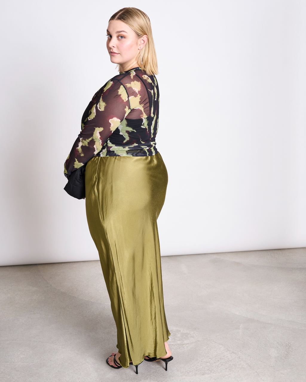 A blonde woman stands in a studio, looking over her shoulder. She wears the jan-n-june MESH LONGSLEEVE DORA MOSSFIRE top with a shiny olive-green skirt and black heeled sandals against a light background.
