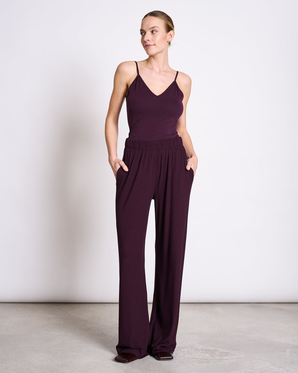 A woman stands against a white background, wearing the jan-n-june TOP MATILDA CHERRY LACQUER GOTS—a sleeveless, dark purple V-neck jersey top—and matching wide-leg pants, with her hands in her pockets and hair pulled back.