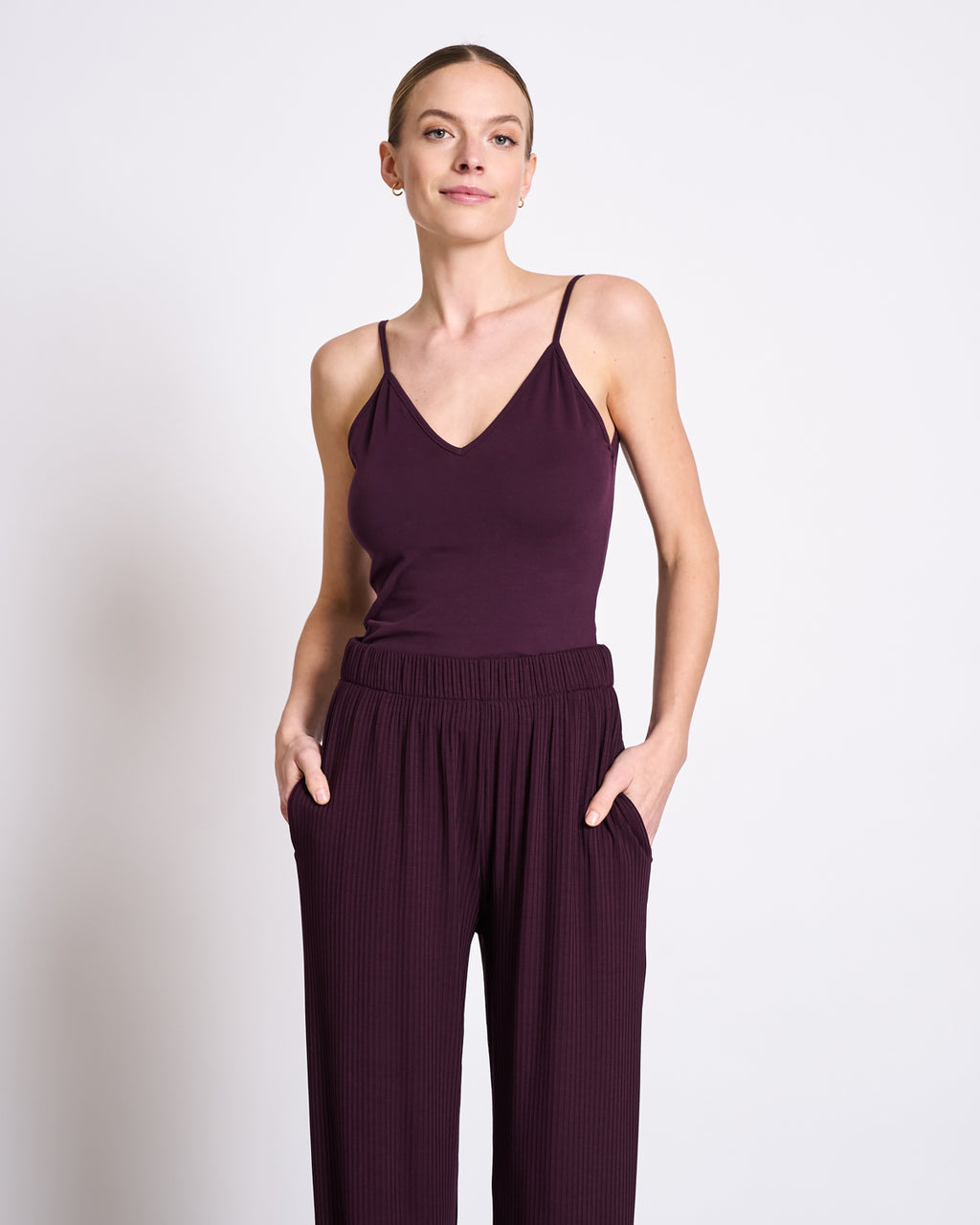 A woman smiles softly at the camera, standing against a white background in the jan-n-june TOP MATILDA CHERRY LACQUER GOTS— a sleeveless deep purple V-neck jersey top—paired with matching loose-fitting pants and her hands in her pockets.