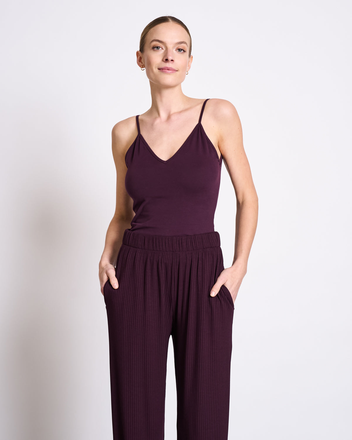A woman smiles softly at the camera, standing against a white background in the jan-n-june TOP MATILDA CHERRY LACQUER GOTS— a sleeveless deep purple V-neck jersey top—paired with matching loose-fitting pants and her hands in her pockets.