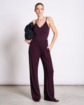 A woman stands against a plain background wearing the jan-n-june TOP MATILDA CHERRY LACQUER GOTS, a sleeveless deep purple V-neck jersey top, with matching wide-leg pants. She holds a black jacket over one shoulder and has slicked-back hair.