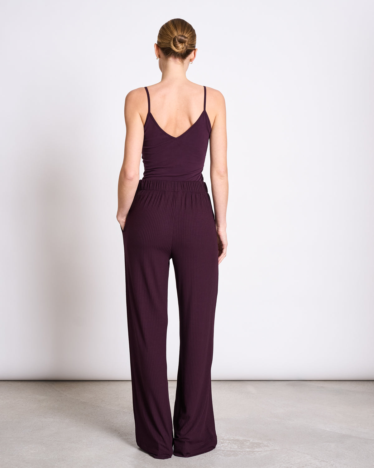 A woman with blonde hair in a low bun stands with her back to the camera, wearing the jan-n-june TOP MATILDA CHERRY LACQUER GOTS, a sleeveless V-neck top, against a plain light background.