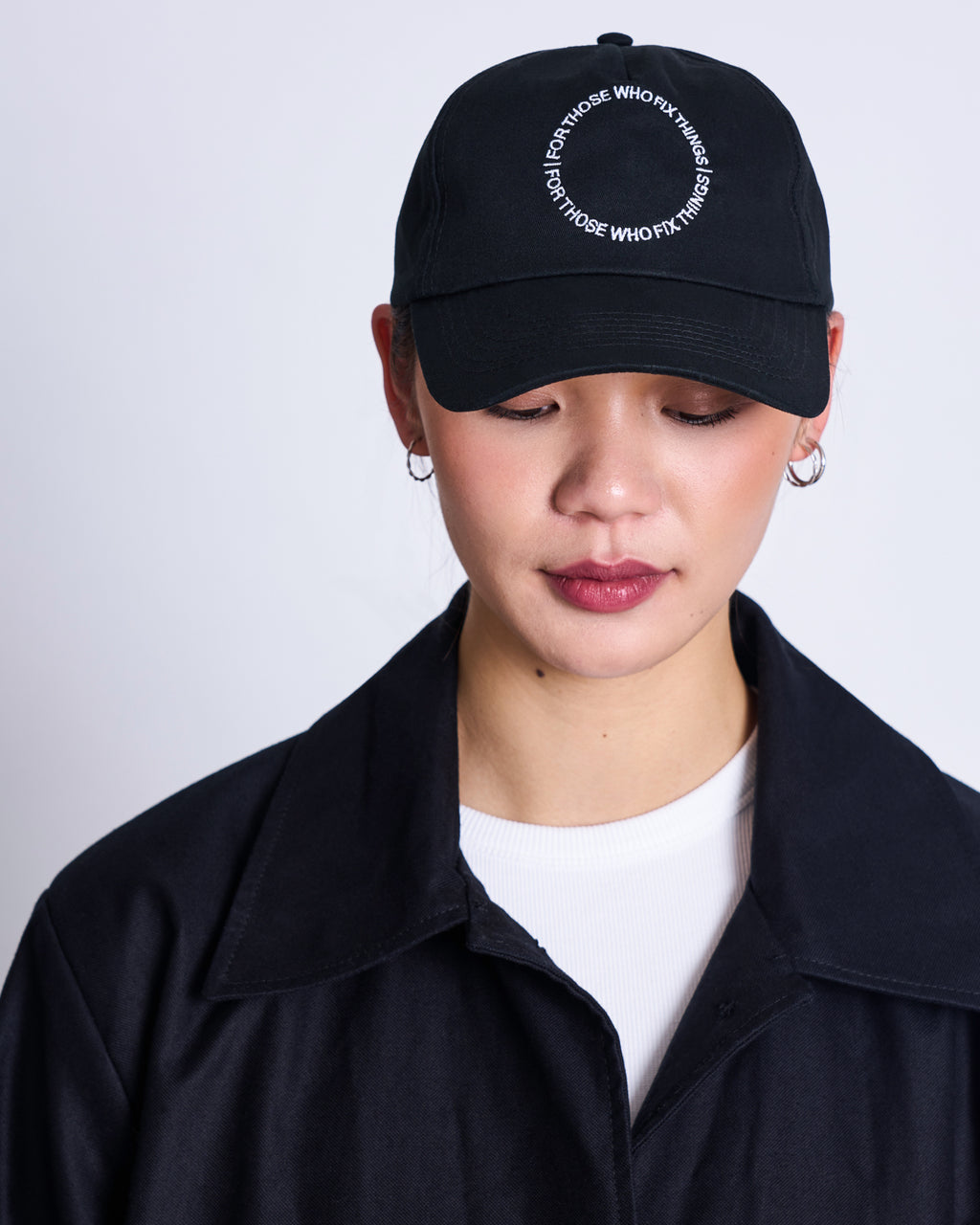 A person wearing the jan-n-june CAP FOR THOSE WHO FIX THINGS BLACK GOTS, featuring circular white text, and a dark jacket over a white shirt looks downward against a plain light background.