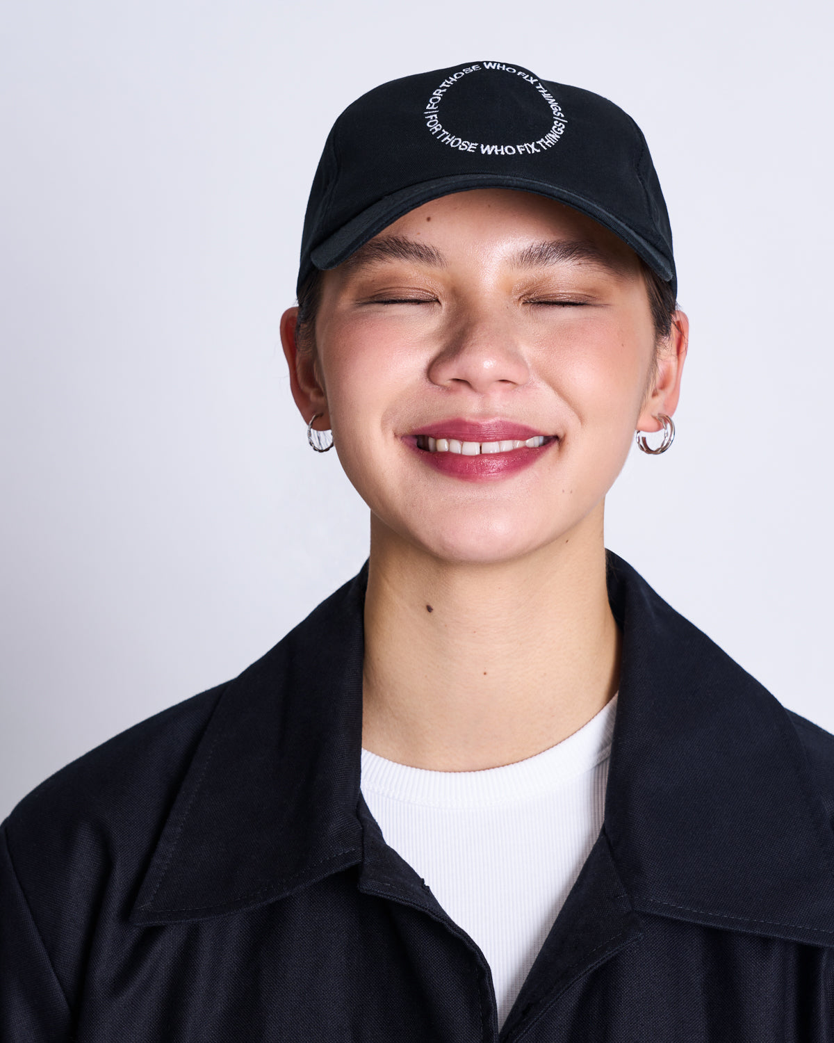 A person with short hair smiles with closed eyes against a plain light background, wearing jan-n-junes black GOTS CAP FOR THOSE WHO FIX THINGS, silver hoop earrings, a black jacket, and a white top.