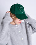 A person wearing a textured gray button-up sweater adjusts the jan-n-june CAP FOR THOSE WHO FIX THINGS BOTTLE GREEN GOTS, its white circular text visible. The cap’s brim and their hands partially obscure their face. The background is plain white.