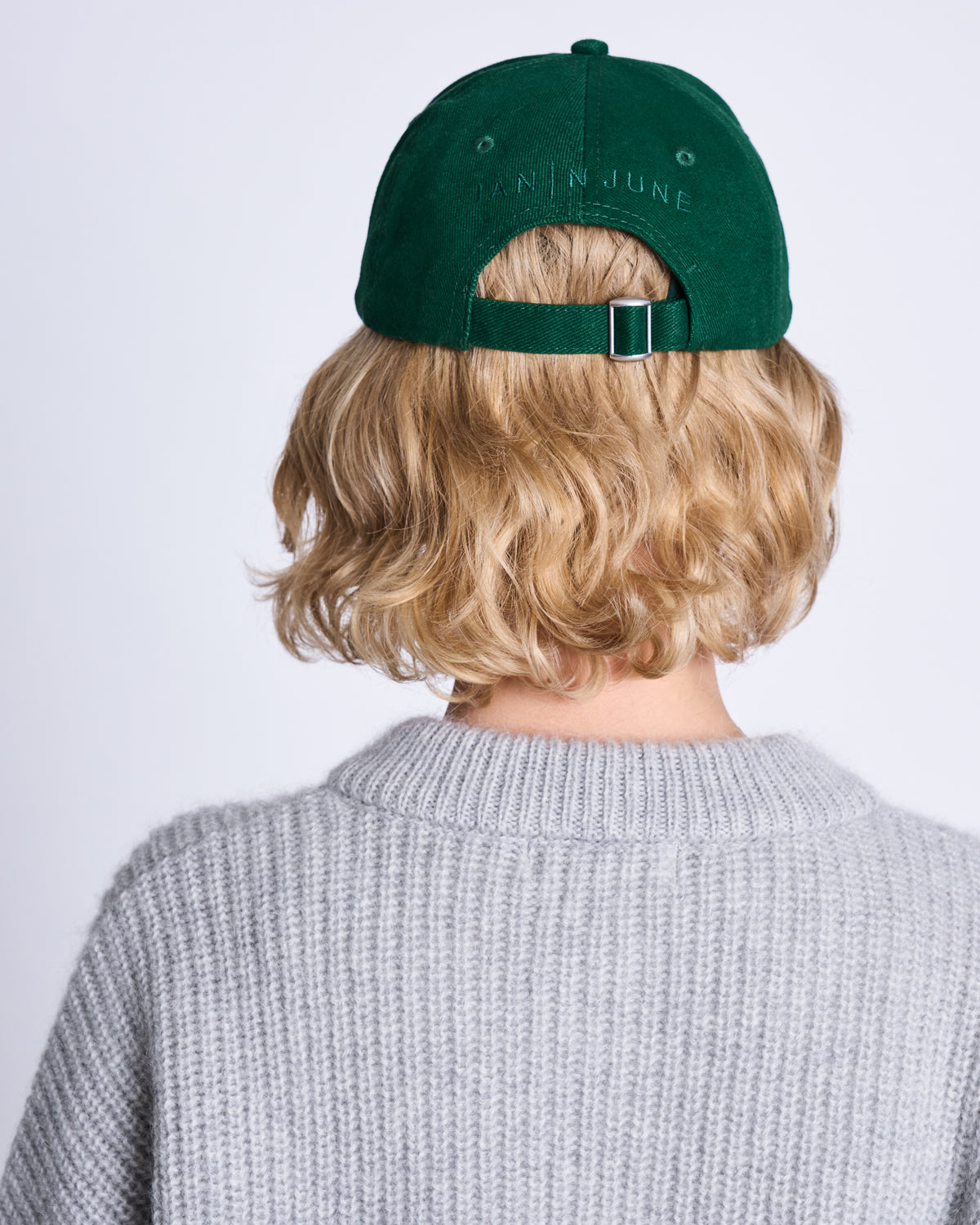 A person with short blonde hair wears the jan-n-june CAP FOR THOSE WHO FIX THINGS BOTTLE GREEN GOTS and a light gray knit sweater, shown from behind against a plain white background.
