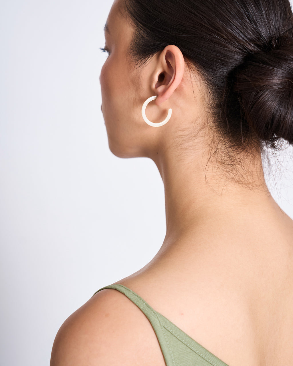 A woman with dark hair in a bun wears jan-n-june MINI HOOPS IVORY acetate earrings and a green sleeveless top, seen from the side and slightly behind against a plain background.